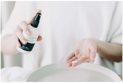 A person sprays lotion onto their hand from a small bottle, with a white background and a gray bowl nearby.