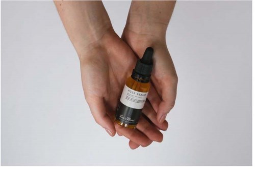 Two hands hold a small brown bottle with a black dropper cap, labeled with skincare or essential oil product details.