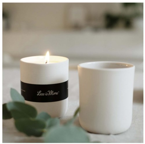 A lit candle with a black label 'Less is More', and an empty white cup on a table with green leaves.
