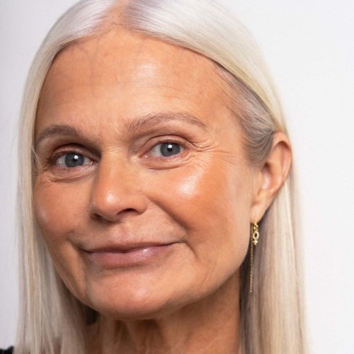 Een oudere vrouw met lang wit haar, lichte huid en blauwe ogen, glimlacht zacht. Ze draagt oorbellen en heeft een rustige uitstraling.