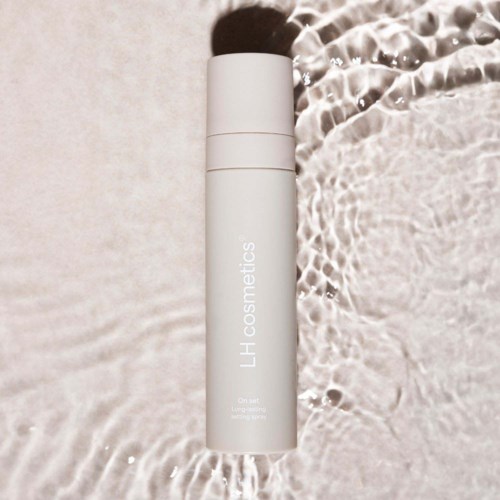 En vit cylinderformad flaska med svart lock på sandytan nära vattnet, märkt "LH cosmetics."