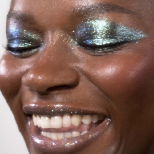 Närbild av en leende person med glittrig makeup på ögonlock och läppar, visar tänder och glad min.