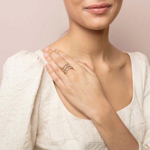 Eine Frau berührt ihre Schulter mit einem Ring am Finger und trägt eine hellfarbene Bluse. Der Hintergrund ist schlicht.