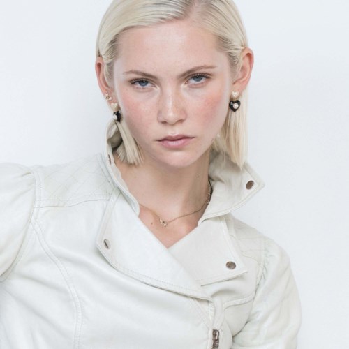 En kvinna med platinablont hår i vit jacka och svarta hjärtformade örhängen, tittar självsäkert mot kameran.