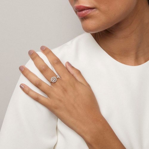 A woman wears a white top and a ring on her finger, touching her shoulder with her hand.