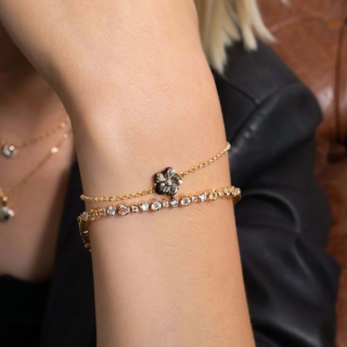 Twee gouden armbanden, een met een bloemhanger en de andere met heldere edelstenen, gedragen op iemands pols.