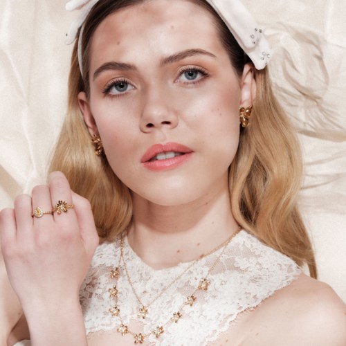 Een jonge vrouw met blond haar draagt elegante sieraden en een kanten jurk, ligt op satijn.