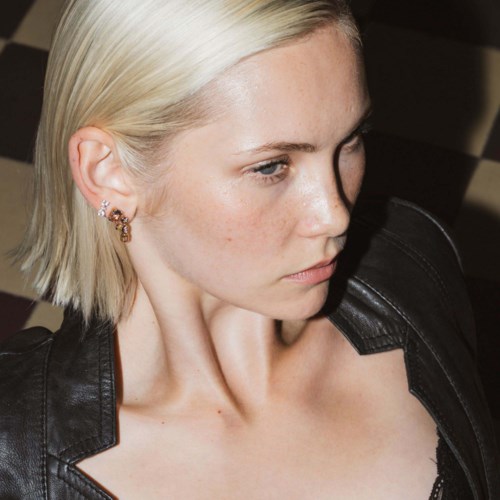 En kvinna med kort blont hår, bär en svart skinnjacka och örhängen, tittar förbi med ett allvarligt ansiktsuttryck.