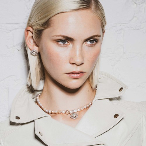 Een vrouw met blond haar en lichte make-up draagt een parelketting, oorbellen en een beige jas tegen een witte bakstenen muur.