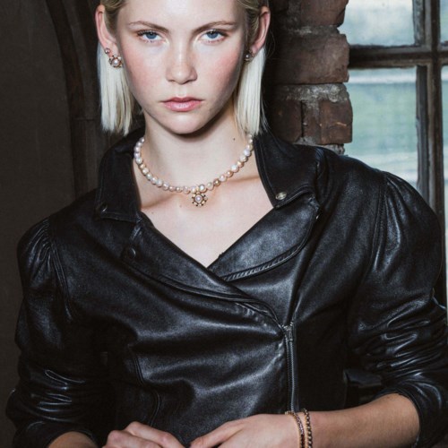 Jonge vrouw met kort blond haar, zwart leren jas en parelketting binnen.
