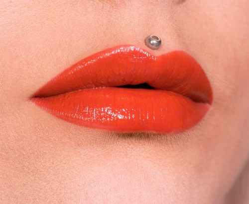 Läppar med stark orange läppstift och en näspiercing ovanför. Huden ser slät ut och bilden är en närbild.