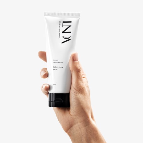En hånd holder en hvit tube med svart hette, merket 'INDIA'. Det er et hudpleieprodukt, en rensende balm.
