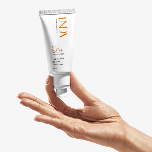 En hånd holder en hvit tube med ansikts solkrem med SPF 50+ mot en hvit bakgrunn.