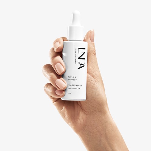 En hånd holder en hvit flaske med pumpe, merket 'INDIA,' inneholder et serum for hårbeskyttelse.