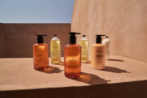 Fem flasker shampoo og balsam er arrangeret på en overflade i klart sollys, der kaster skygger. Klare etiketter er synlige.
