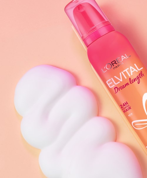 En rosa flaska med Loreal Elvital Dream Length hårprodukt och vitt skum på en persikofärgad bakgrund.