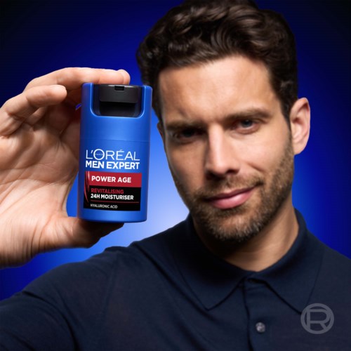 En man håller en blå flaska Loreal Men Expert Power Age återfuktare nära ansiktet mot en mörkblå bakgrund.