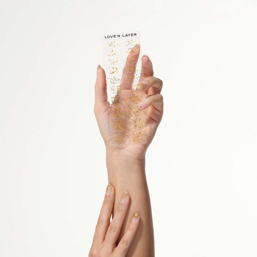 En hand med dekorerade naglar håller ett klart ark med titeln "Love'n Layer" och guldglitter på envit bakgrund.
