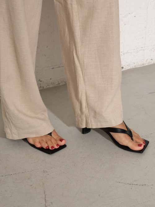 Persoon draagt beige broek en zwarte slippers. Een klein deel van hun voeten met rode nagellak is zichtbaar.