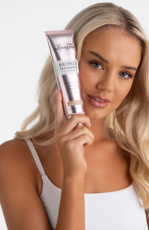 A woman with long blonde hair holds a tube of bronzer shimmer product close to her face.
