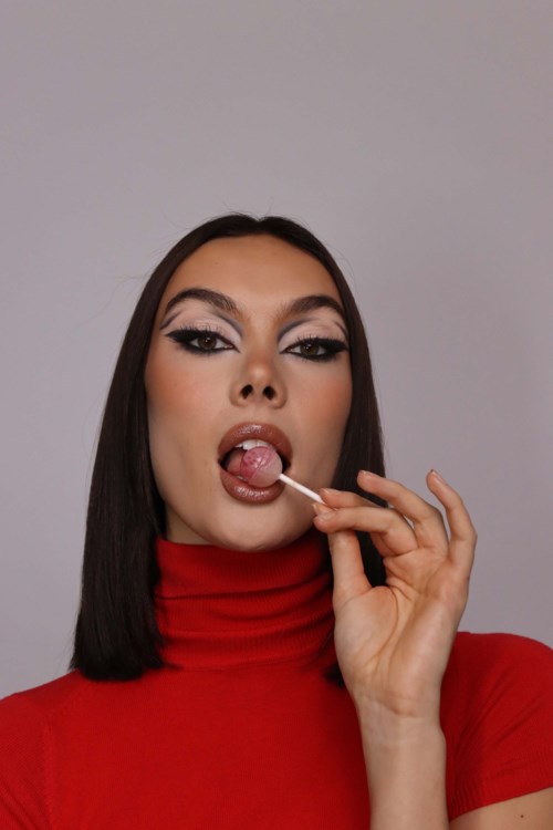 A woman in a red turtleneck dress is licking a lollipop, with makeup and black hair, against a plain background.