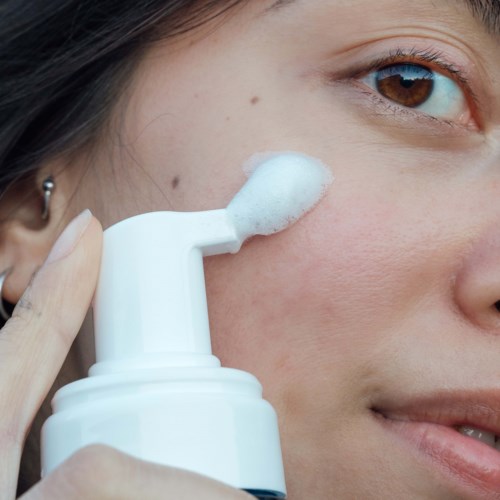A person applies foamy face cleanser under the eye area. Close-up of face and hand with skincare tool. Clear and simple.