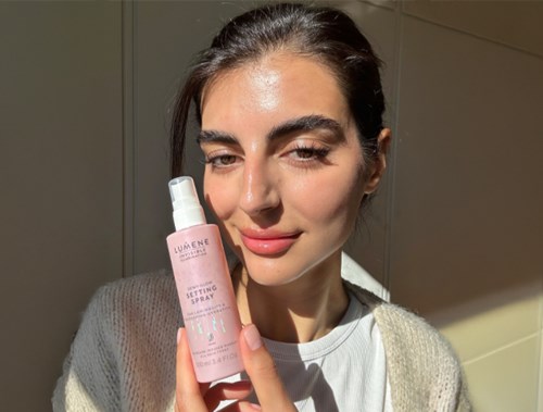 En kvinne holder en rosa flaske med setting spray, smilende med sollys på ansiktet.