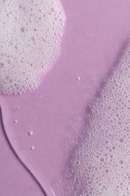 Pink skum med bobler på lilla baggrund, glat og skummende tekstur. Det ligner sæbe- eller badebadeskum.
