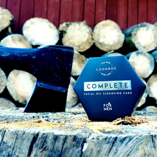 En blå sekskantet boks merket "Complete Facial Oil Cleansing Cake" for menn, med trekker i bakgrunnen og et svart objekt i nærheten.