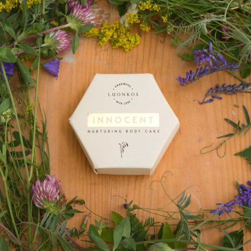 Eine sechseckige Box mit 'Innocent Nurturing Body Cake' beschriftet, umgeben von bunten Blumen auf Holztisch.