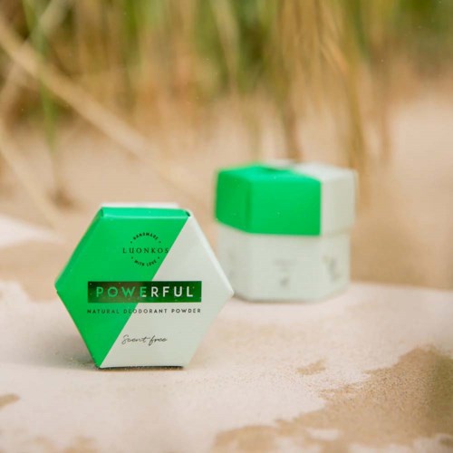 A green and white container of natural deodorant powder labeled 'Powerful' on a sandy surface with blurred plants in the background.