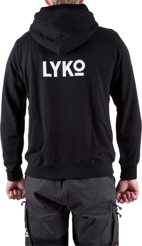 En person bär en svart hoodie med vitt "LYKO"-text på ryggen, tillsammans med mörka byxor i en avslappnad miljö.