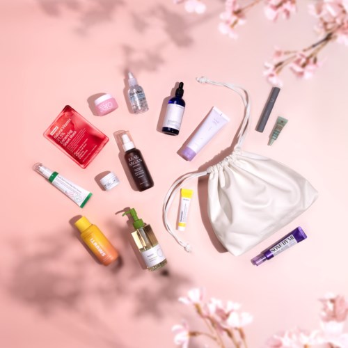 Verschillende skincare- en beautyproducten rondom een witte koordtasje op een roze achtergrond met kersenbloesems.