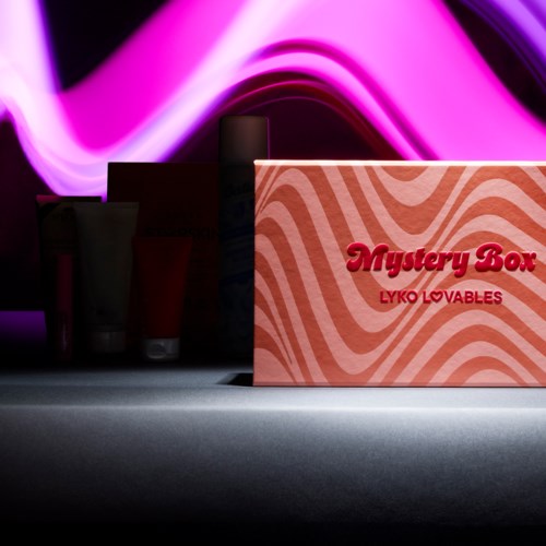 En rosa låda märkt 'Mystery Box' med skönhetsprodukter bakom den på en mörk yta.