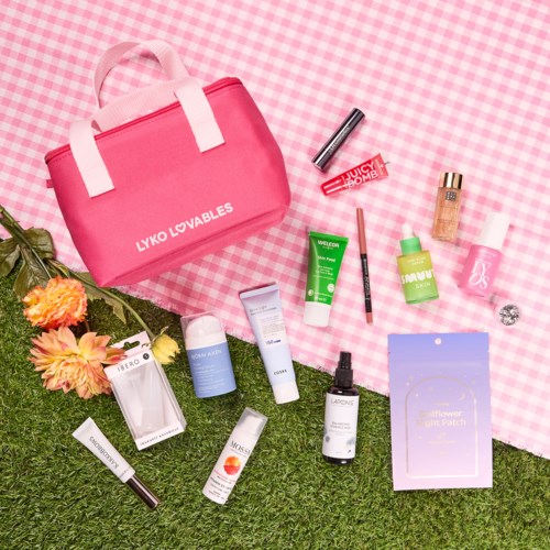 Eine pinke Tasche und verschiedene Hautpflege- und Make-up-Produkte auf Gras und karierter Unterlage.