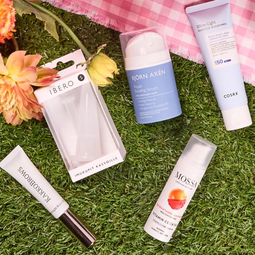 Hautpflegeprodukte und Blumen auf Gras, inklusive Sonnencreme, Serum und Lippenprodukt mit pinken Blumen.