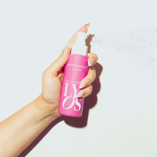 Dłoń trzyma różowy spray z białym tekstem, na białym tle. Butelka ma napis "LIYOS."