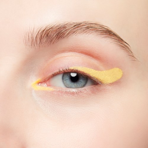 An eye with a yellow eye mask on the eyelid, clear skin, and blonde eyebrow hairs.