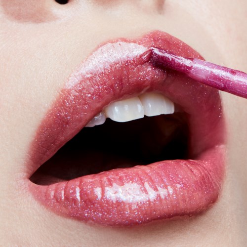 En person påfører lipgloss med en liten børste, munnen er litt åpen, viser tenner og lyse rosa leppestift.
