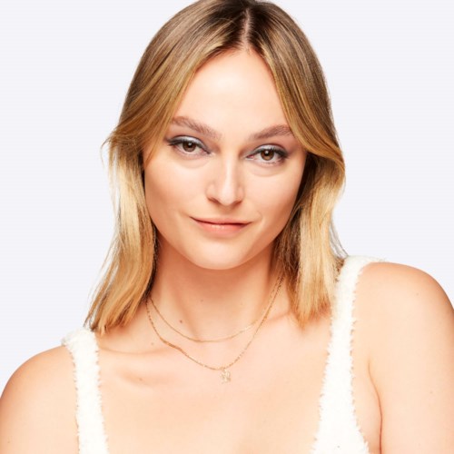 En ung kvinde med blondt hår, let makeup og et blidt smil, iført hvid top og lagdelte halskæder.
