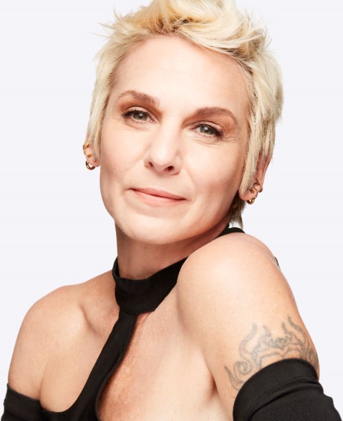 En kvinna med kort blont hår som ler mjukt, bär en svart topp, visar en tatuering på axeln.