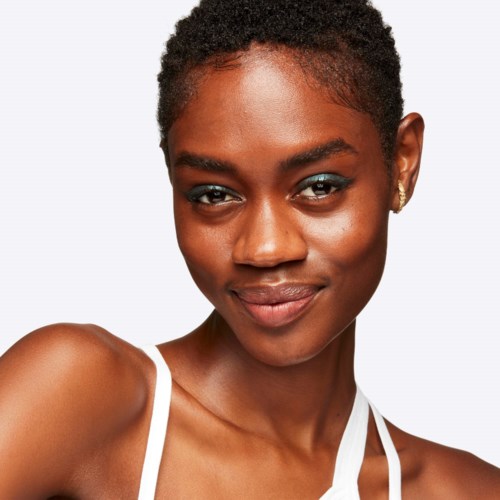 A smiling young woman with short, curly hair and dark skin, wearing a white top, looks directly at the camera.