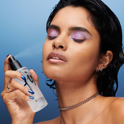 Eine Frau mit kurzen dunklen Haaren und lila Make-up verwendet eine Sprühflasche, mit geschlossenen Augen und einem ruhigen Ausdruck.