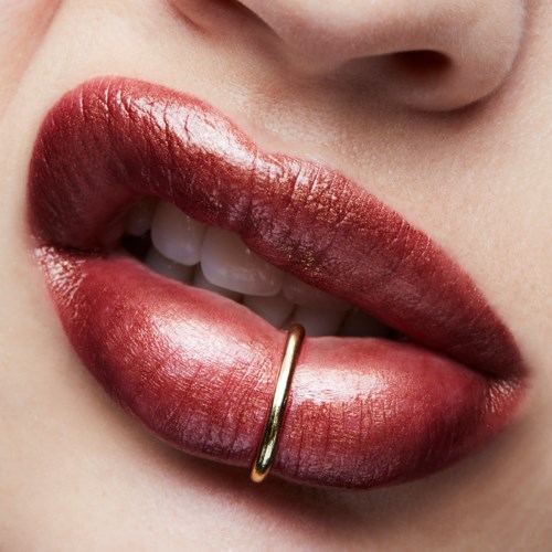 Close-up van lippen met rode lippenstift, een kleine gouden ring op de onderlip en zichtbaar gebit.