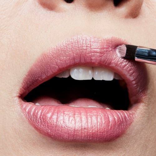 Iemand brengt lichtroze lippenstift aan op lippen, met lichte tanden zichtbaar. Close-up op de mond en lippenstiftborstel.