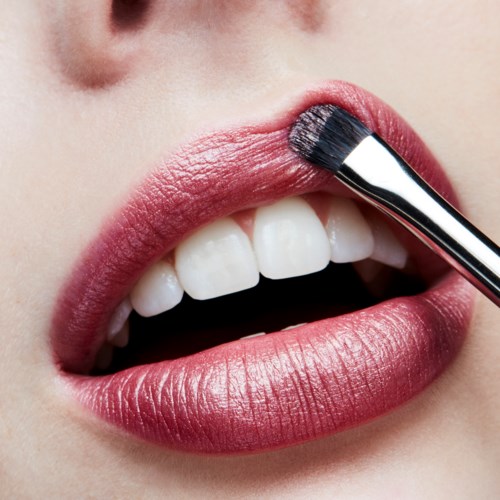Een persoon brengt lippenstift aan met een kwast op de lippen, close-up van mond en tanden.