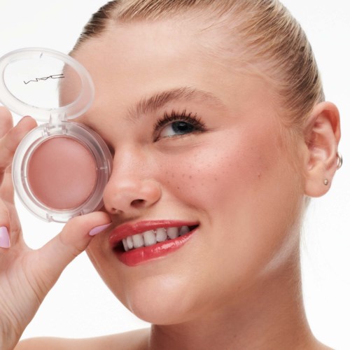 A woman holds a pink makeup compact near her eye, smiling. She has blonde hair and bright skin with freckles.