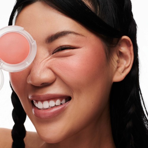 A smiling woman with braided black hair holds a pink blush compact near her face.