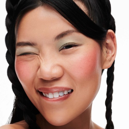A woman with dark hair in braids, winking with a big smile showing teeth, and wearing makeup.