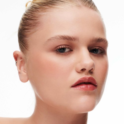 En kvinde med glat hud, let makeup og blonde hår bundet tilbage, ser selvsikker ud mod kameraet.